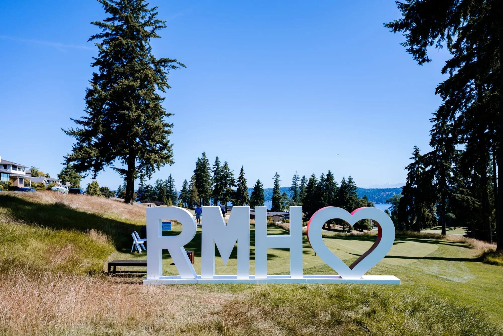 Giant white RMH letters and a heart, a Ronald McDonald House sign, on a golf course framed by evergreens